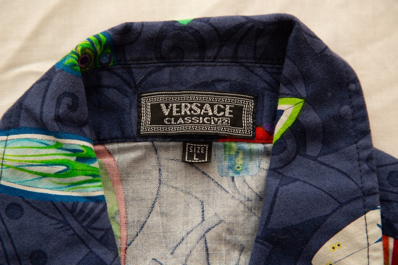 May include: Close-up of a navy blue button-down shirt with a colorful, abstract pattern. The shirt collar features a label with the "VERSACE CLASSIC V2" logo and a size "L" tag.