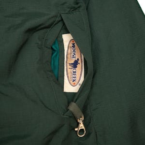 May include: Dark green jacket with a pocket detail. The pocket contains a tag with the text "MOOSE CREEK CLOTHING" and a graphic of a person with a moose. A gold-colored clasp is attached to the pocket.