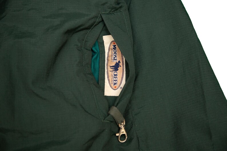 May include: Dark green jacket with a pocket detail. The pocket contains a tag with the text "MOOSE CREEK CLOTHING" and a graphic of a person with a moose. A gold-colored clasp is attached to the pocket.