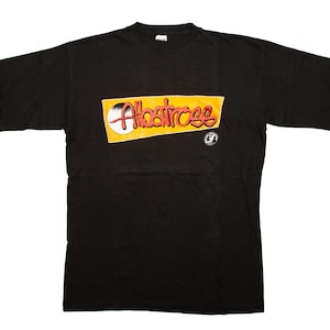 Könnte beinhalten: Schwarzes T-Shirt mit dem Wort "Albatross" in roter Schrift auf einem gelben Rechteck. Ein weißer Kreis und eine kleine Grafik befinden sich rechts vom Text. Das Shirt hat einen Rundhalsausschnitt.
