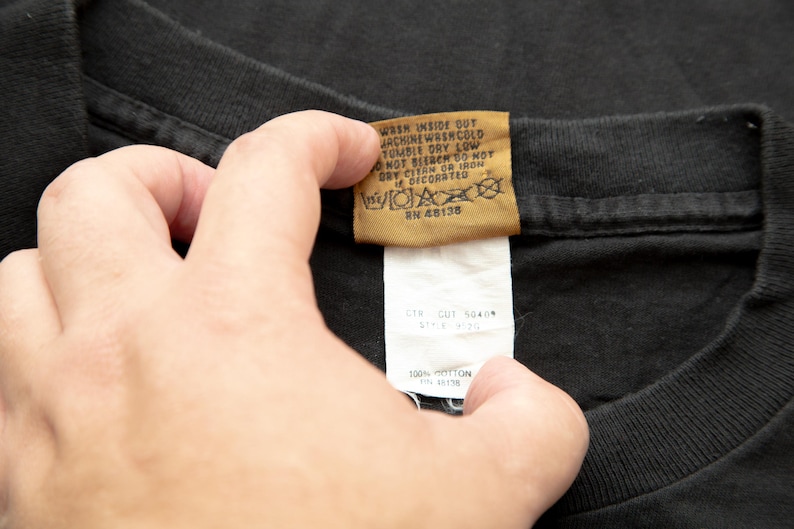 K&ouml;nnte beinhalten: Ein schwarzes T-Shirt mit einem braunen Pflegeetikett, auf dem steht "Wash inside out, machine wash cold, tumble dry low, do not bleach, do not iron or dry clean, not decorated, RN 48138". Das Etikett ist an einem wei&szlig;en Etikett befestigt, auf dem "CTR Cut 5040" und "100% Cotton" steht.