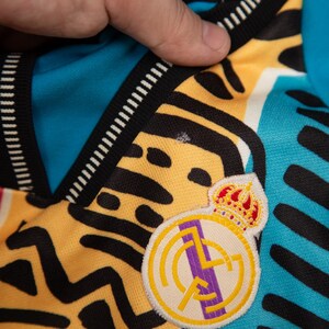 May include: Close-up of a jersey with a colorful design. The jersey features a yellow and black pattern, with a blue and red section. The Real Madrid crest is visible on the jersey.