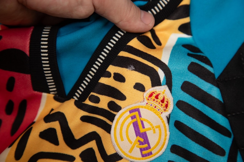 May include: Close-up of a jersey with a colorful design. The jersey features a yellow and black pattern, with a blue and red section. The Real Madrid crest is visible on the jersey.