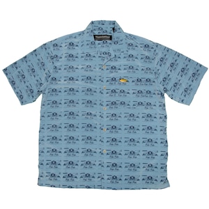 Op de afbeelding: Lichtblauwe shirt met korte mouwen en een herhalend patroon van kleine donkerblauwe ontwerpen. Het shirt heeft een kraag en een klein label aan de bovenkant. Het shirt is gemaakt van een lichte stof.