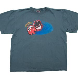 Op de afbeelding: Een donkerblauw t-shirt met een afbeelding van twee rode en blauwe cartoon gezichten met tanden. De tekst "Spitfire" staat boven de gezichten.