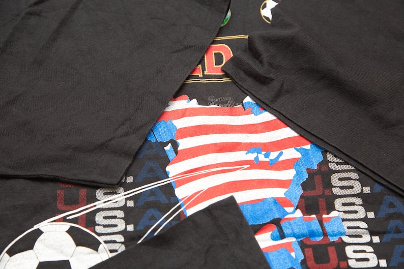 K&ouml;nnte beinhalten: Schwarzes T-Shirt mit einem Grafikdruck eines Fu&szlig;balls, den Worten "U.S.A." und einer stilisierten Karte der Vereinigten Staaten mit roten und wei&szlig;en Streifen.