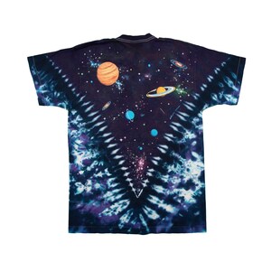 May include: A tie-dye t-shirt featuring a vibrant space-themed design. The shirt showcases planets, galaxies, and stars against a dark blue and purple background. The design is centered on the back of the shirt.