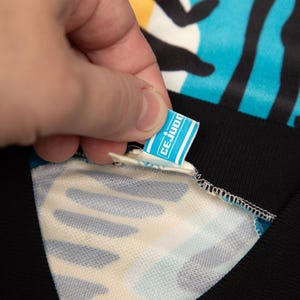 May include: Close-up of a blue and black patterned garment with a black collar. A small tag with the text "CE JUDO" is being held up by a hand. The garment has a white and blue pattern.