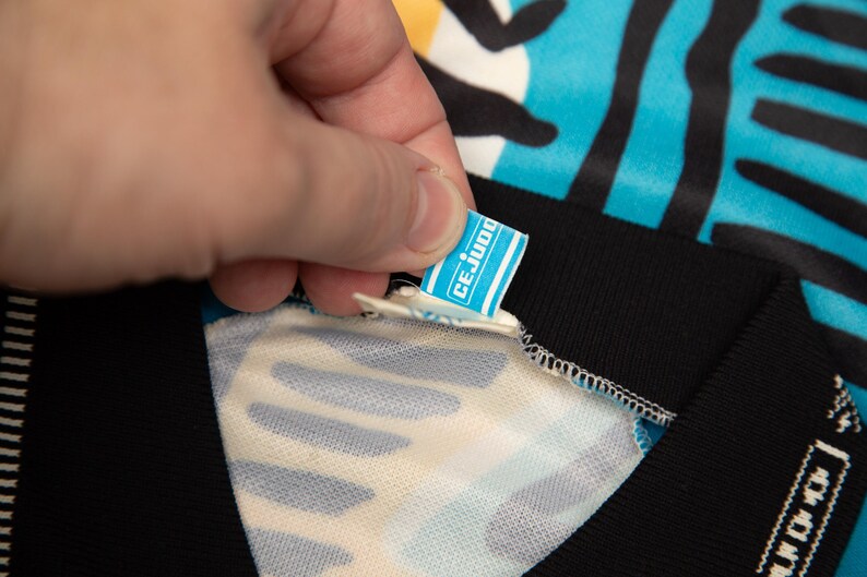 May include: Close-up of a blue and black patterned garment with a black collar. A small tag with the text "CE JUDO" is being held up by a hand. The garment has a white and blue pattern.