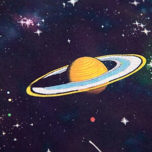 May include: A close-up of a dark blue fabric featuring a vibrant illustration of a planet with yellow rings and a textured surface, set against a starry space background. The design includes white stars and colorful nebulas.
