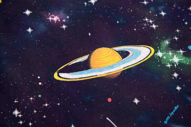 May include: A close-up of a dark blue fabric featuring a vibrant illustration of a planet with yellow rings and a textured surface, set against a starry space background. The design includes white stars and colorful nebulas.