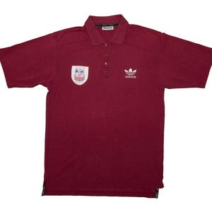 Pode incluir: Polo bordô de manga curta. A camisa apresenta um logotipo Adidas branco no peito direito e um patch no peito esquerdo. Possui gola e carcela de três botões.
