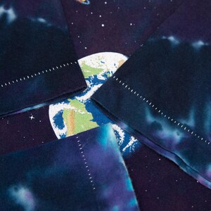 May include: Close-up of a tie-dye t-shirt featuring a space-themed design. The shirt displays a vibrant Earth graphic, surrounded by stars and planets. The sleeves have a dark blue and purple tie-dye pattern with white stitching.