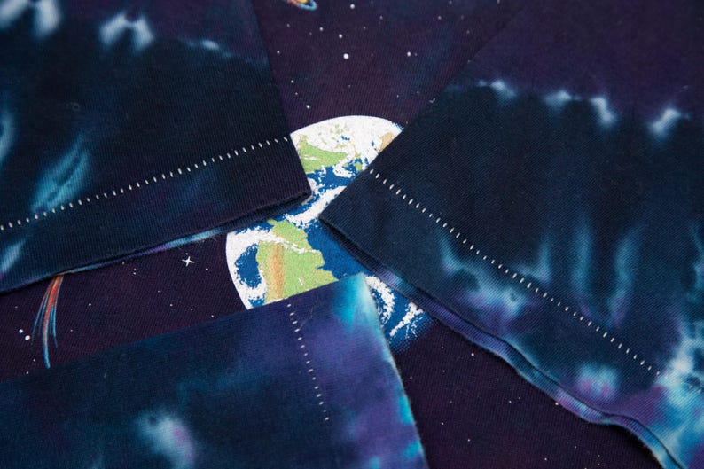May include: Close-up of a tie-dye t-shirt featuring a space-themed design. The shirt displays a vibrant Earth graphic, surrounded by stars and planets. The sleeves have a dark blue and purple tie-dye pattern with white stitching.