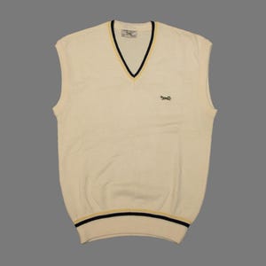 Pode incluir: Camisola sem mangas creme com decote em V e um pequeno logótipo bordado. A gola e a bainha inferior apresentam riscas pretas e amarelas. Um estilo clássico e preppy.