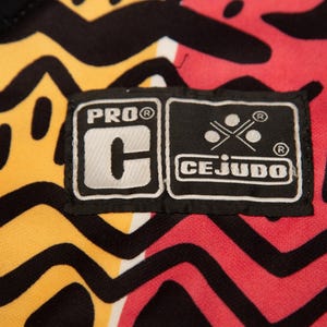May include: Close-up of a fabric with a vibrant pattern in yellow, red, and black. The design features bold, abstract shapes and lines. A black label with white text reads "PRO C" and "CEJUDO".