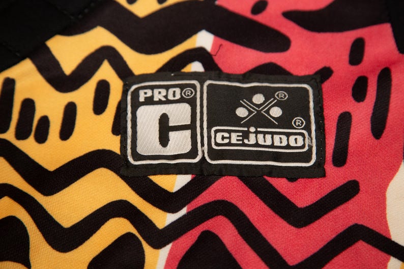May include: Close-up of a fabric with a vibrant pattern in yellow, red, and black. The design features bold, abstract shapes and lines. A black label with white text reads "PRO C" and "CEJUDO".