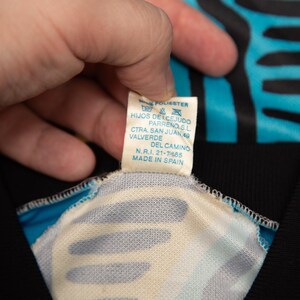 May include: Close-up of a tag from a garment, held by a hand. The tag is white with blue text, and reads "100% POLIESTER" and "MADE IN SPAIN". The garment has a black and turquoise pattern.