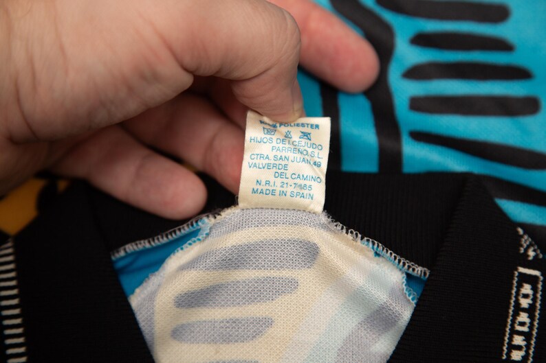 May include: Close-up of a tag from a garment, held by a hand. The tag is white with blue text, and reads "100% POLIESTER" and "MADE IN SPAIN". The garment has a black and turquoise pattern.
