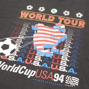 K&ouml;nnte beinhalten: Schwarzes T-Shirt mit einem Grafikdruck eines Fu&szlig;balls, der eine Karte der Vereinigten Staaten tritt. Die Grafik enth&auml;lt auch den Text "World Tour" und "World Cup USA 94".