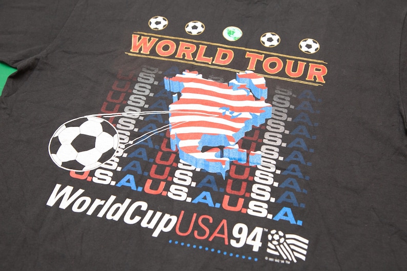 K&ouml;nnte beinhalten: Schwarzes T-Shirt mit einem Grafikdruck eines Fu&szlig;balls, der eine Karte der Vereinigten Staaten tritt. Die Grafik enth&auml;lt auch den Text "World Tour" und "World Cup USA 94".