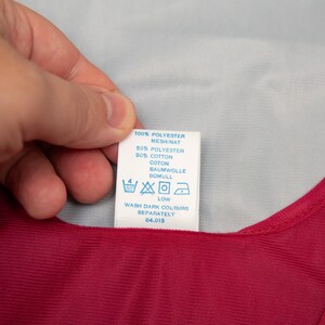 May include: A white clothing label with black text and washing symbols is held over a red garment. The label reads: 100% POLYESTER, 50% POLYESTER, 50% COTTON, WASH DARK COLOURS SEPARATELY.