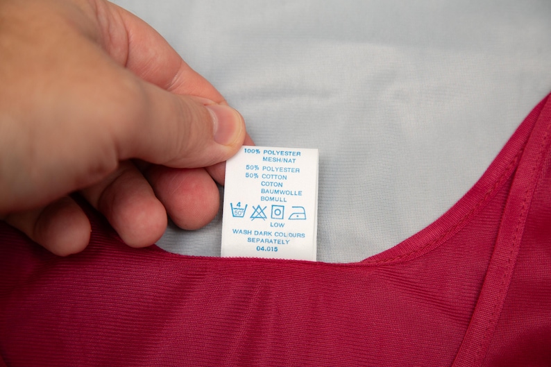 May include: A white clothing label with black text and washing symbols is held over a red garment. The label reads: 100% POLYESTER, 50% POLYESTER, 50% COTTON, WASH DARK COLOURS SEPARATELY.