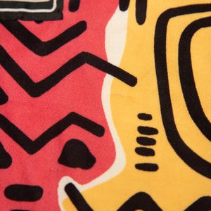 May include: Close-up of a fabric with a vibrant abstract design. The pattern features bold black shapes on a red and yellow background. The design includes zigzags, triangles, and spiral elements, creating a striking visual.