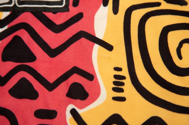 May include: Close-up of a fabric with a vibrant abstract design. The pattern features bold black shapes on a red and yellow background. The design includes zigzags, triangles, and spiral elements, creating a striking visual.