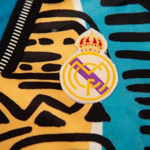 May include: Close-up of a jersey with a Real Madrid patch. The jersey has a yellow and black pattern on the left side, and a blue and black pattern on the right side. The patch features a crown and the Real Madrid logo.