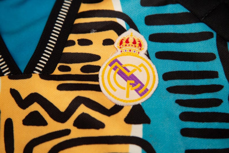 May include: Close-up of a jersey with a Real Madrid patch. The jersey has a yellow and black pattern on the left side, and a blue and black pattern on the right side. The patch features a crown and the Real Madrid logo.