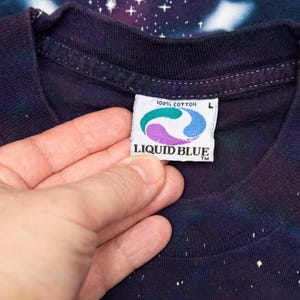 May include: Close-up of a dark blue t-shirt with a galaxy print. The shirt's tag reads "100% Cotton" and features the Liquid Blue logo. The shirt is size L.