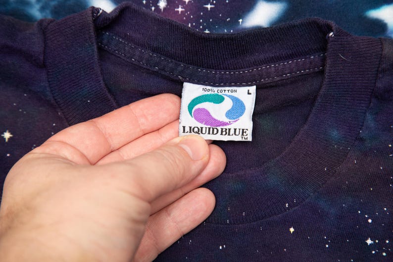 May include: Close-up of a dark blue t-shirt with a galaxy print. The shirt's tag reads "100% Cotton" and features the Liquid Blue logo. The shirt is size L.