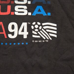 K&ouml;nnte beinhalten: Schwarzes T-Shirt mit wei&szlig;em Text, der "USA 94" lautet, und einer Fu&szlig;ballgrafik. Der Text "USA" ist in Rot, Wei&szlig; und Blau. Der Text "94" ist in Wei&szlig;. Der Fu&szlig;ball ist in Wei&szlig; und Schwarz. Der Text "1991 WC '94 TM" ist in Wei&szlig;.
