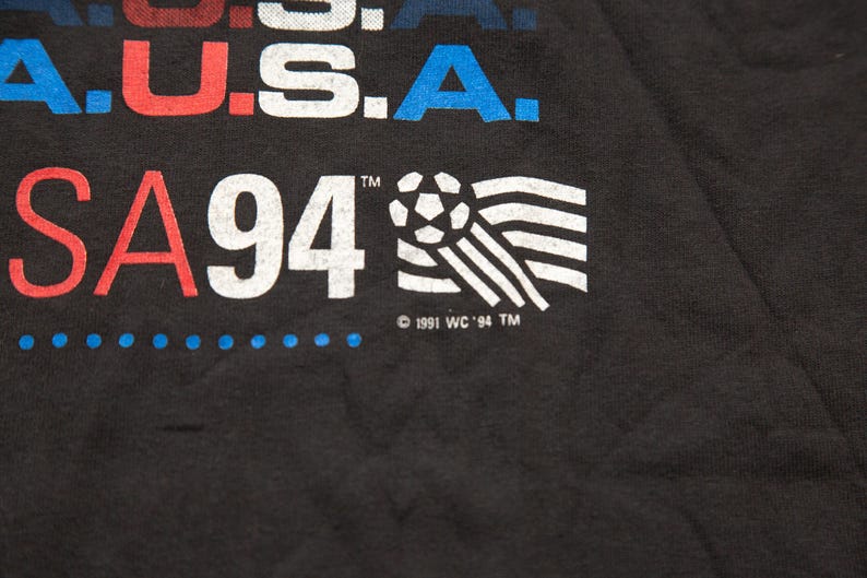 K&ouml;nnte beinhalten: Schwarzes T-Shirt mit wei&szlig;em Text, der "USA 94" lautet, und einer Fu&szlig;ballgrafik. Der Text "USA" ist in Rot, Wei&szlig; und Blau. Der Text "94" ist in Wei&szlig;. Der Fu&szlig;ball ist in Wei&szlig; und Schwarz. Der Text "1991 WC '94 TM" ist in Wei&szlig;.