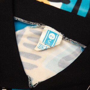May include: Close-up of a colorful garment with a black collar and trim. The fabric features a vibrant pattern of blue, yellow, and pink. A small tag with the number "2" is visible.
