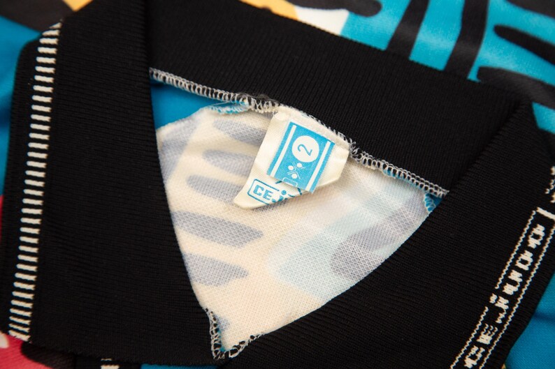 May include: Close-up of a colorful garment with a black collar and trim. The fabric features a vibrant pattern of blue, yellow, and pink. A small tag with the number "2" is visible.