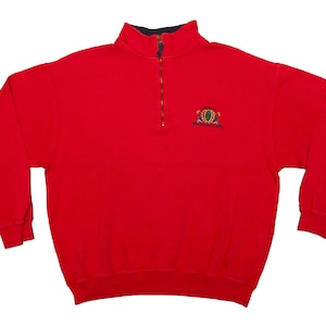 Può includere: Una felpa rosso brillante con collo a zip a un quarto e maniche lunghe. Il colletto è blu navy. Un piccolo stemma è ricamato sul petto sinistro. La felpa è di colore pieno e sembra essere realizzata in un materiale morbido.