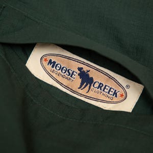 May include: Close-up of a dark green garment with a pocket. A rectangular label with the text "MOOSE CREEK CLOTHING" and a moose silhouette is visible inside the pocket. The label has a tan background with red and blue accents.