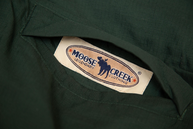 May include: Close-up of a dark green garment with a pocket. A rectangular label with the text "MOOSE CREEK CLOTHING" and a moose silhouette is visible inside the pocket. The label has a tan background with red and blue accents.