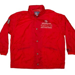 Könnte beinhalten: Rote San Francisco 49ers Jacke mit dem Teamlogo und dem Aufdruck "49ers" auf der Vorderseite. Die Jacke hat einen Knopfverschluss, lange Ärmel und einen Kragen. Das Logo der National Football Conference ist ebenfalls sichtbar.