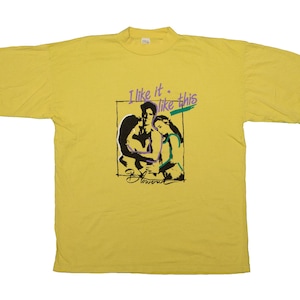 Op de afbeelding: Geel t-shirt met een afbeelding van twee figuren en de tekst "I like it like this". De afbeelding is in zwart, paars en groen. Het shirt heeft een vintage stijl.