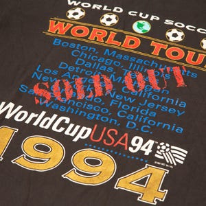 K&ouml;nnte beinhalten: Schwarzes T-Shirt mit einem wei&szlig;en Grafikdruck der World Cup USA Tour 1994. Die Grafik enth&auml;lt den Text "World Cup Soccer" und "World Tour" sowie eine Liste von St&auml;dten in den Vereinigten Staaten.