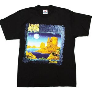 Pode incluir: Camiseta preta com um gráfico de uma paisagem desértica com lua cheia, água e formações rochosas. O texto "Lynyrd Skynyrd" e "Twenty" são visíveis no gráfico. A camiseta tem mangas curtas e gola redonda.