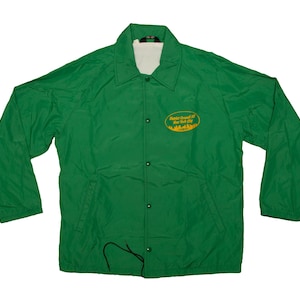 Könnte beinhalten: Eine grüne Nylonjacke mit weißem Innenfutter. Die Jacke hat eine Knopfleiste, lange Ärmel und einen Kragen. Ein ovales Abzeichen mit gelbem Text und Stadtlandschaft befindet sich auf der linken Brust. Die Jacke hat eine einfarbige grüne Farbe.
