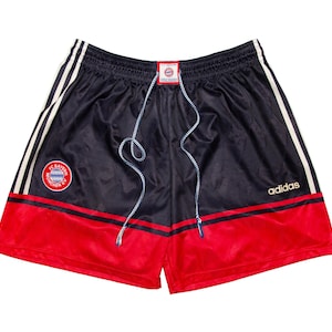 Könnte beinhalten: Marineblaue und rote Adidas-Shorts mit weißen Streifen an den Seiten. Die Shorts zeigen das FC Bayern München-Logo und einen Kordelzug in der Taille. Das Adidas-Logo befindet sich am rechten Bein.