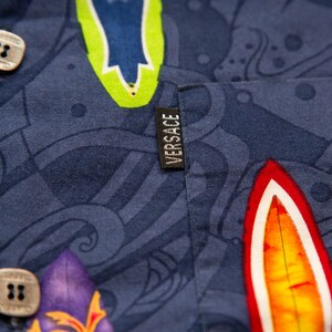 May include: Close-up of a navy blue button-down shirt with a colorful surfboard pattern. The shirt features a small black tag with the brand name "VERSACE" in white. The buttons are square and light-colored.