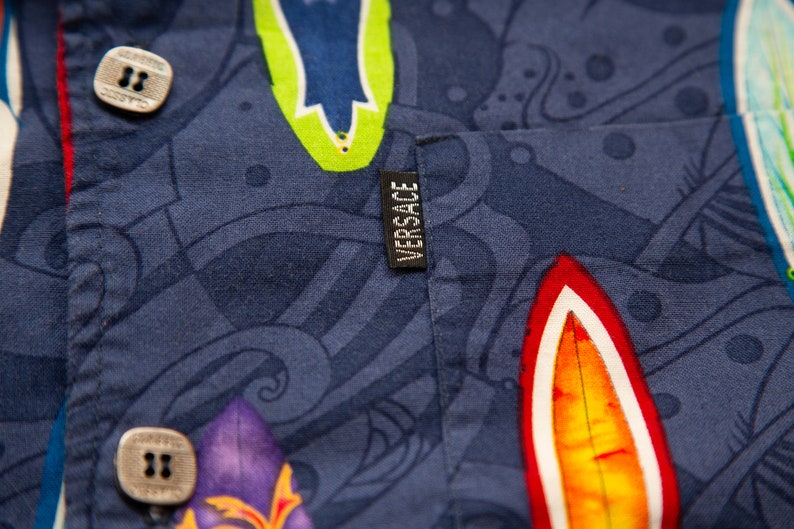 May include: Close-up of a navy blue button-down shirt with a colorful surfboard pattern. The shirt features a small black tag with the brand name "VERSACE" in white. The buttons are square and light-colored.