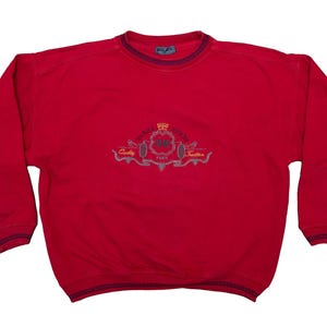 Può includere: Felpa rossa con bordino blu navy intorno al collo e ai polsini. La felpa ha un logo ricamato sul davanti che dice "Champs Elysees" con una corona e altri elementi decorativi.