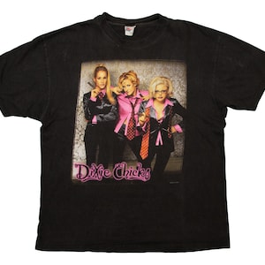 Peut inclure: T-shirt noir avec un graphisme des Dixie Chicks. L'image montre trois femmes en costumes noirs et chemises roses. Le nom du groupe est écrit en rose en bas de l'image.
